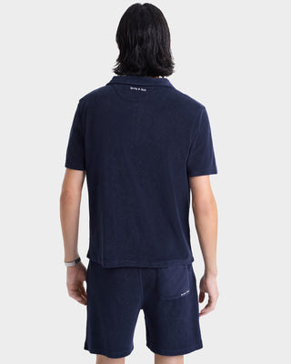 Person with dark hair wearing the Eden Script Terry Polo - Navy/White, styled with matching shorts and shown from the back; a cotton polo crafted in Portugal.