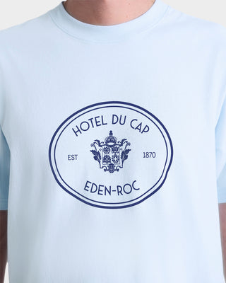 Person wearing the Eden Crest T-Shirt - Ciel/Navy, an oversized 100% cotton light blue tee with HOTEL DU CAP EDEN-ROC and crest printed on the front.
