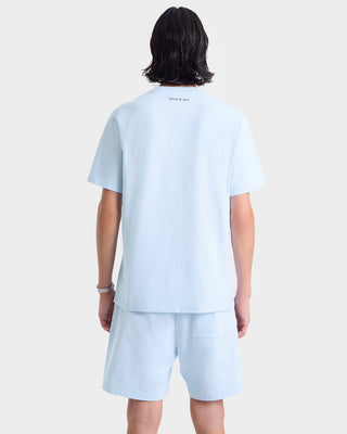 Person with medium-length dark hair, shown from the back, wearing an oversized Eden Crest T-Shirt in Ciel/Navy and shorts.