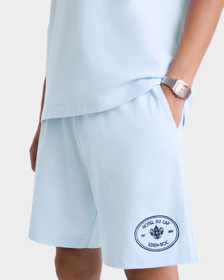 A person wearing the Eden Crest Gym Short in Ciel/Navy, a matching shirt with a Hotel du Cap Eden-Roc patch, and a watch.