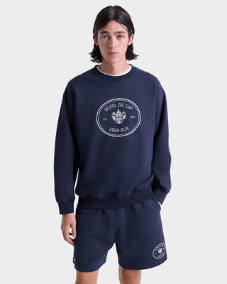Wearing the Eden Crest Crewneck in navy/white and shorts with the Hotel Du Cap Eden-Roc logo, made in Portugal from 100% cotton, person stands against a white background.