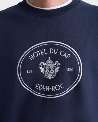 The Eden Crest Crewneck - Navy/White is an oversized navy sweatshirt with HOTEL DU CAP EDEN-ROC EST 1870 and a white crest on the front, made in Portugal from 100% cotton.