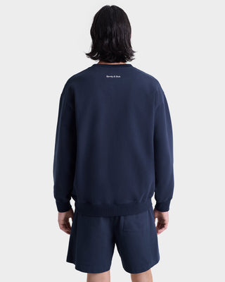 A person with shoulder-length dark hair is shown from the back against a white background, wearing the Eden Crest Crewneck - Navy/White and shorts, made in Portugal from 100% cotton.
