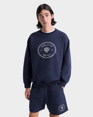 A young person wears the Eden Crest Gym Short in Navy/White, featuring the Hotel du Cap Eden-Roc logo, and stands against a white background.