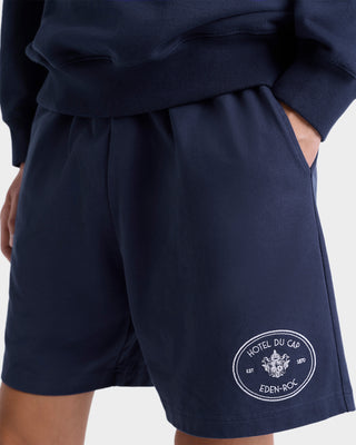 Person wearing Eden Crest Gym Short - Navy/White, featuring the Hotel Du Cap Eden-Roc logo embroidered on the left leg, made in Portugal, with hand in pocket.