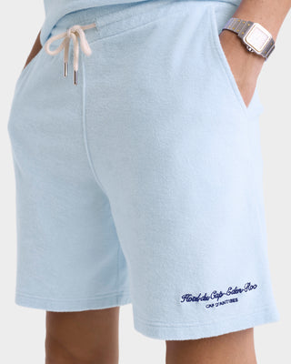 A person wears the Eden Script Terry Short in Ciel/Navy, featuring a light blue hue, visible embroidery near the hem, hands in pockets, a watch, and crafted from 100% cotton for superior comfort and quality.