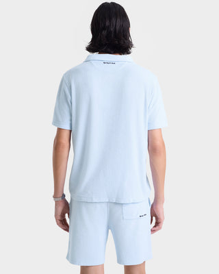 Person facing away, wearing the Eden Script Terry Short in Ciel/Navy—100% cotton, made in Portugal—paired with a matching light blue short-sleeve shirt against a plain background.