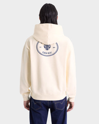 Person seen from behind wearing the Eden Crest Hoodie in Cream/Navy, made in Portugal with an oversized fit, featuring a blue crest and EDEN-ROC text on the back.