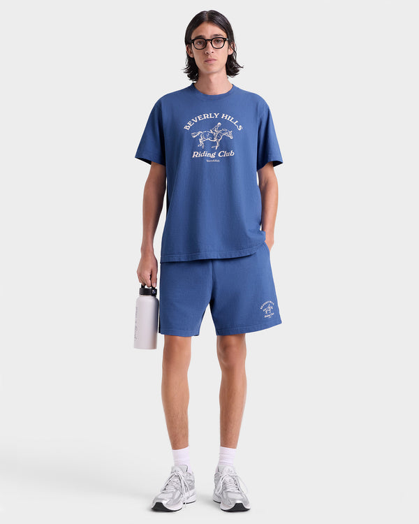 Person wearing a blue t-shirt and shorts with a logo, holding a white bag on a white background