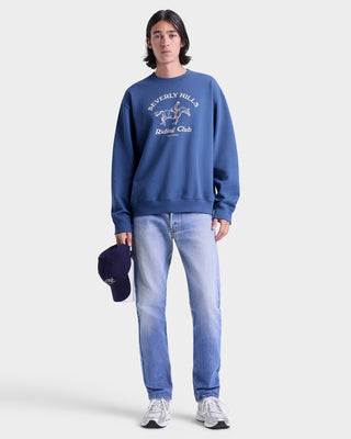 Person wearing a blue sweatshirt with a logo and text, light blue jeans, and a dark cap on a white background