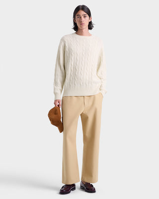 Person wearing a cream sweater and beige pants holding a brown bag on a white background