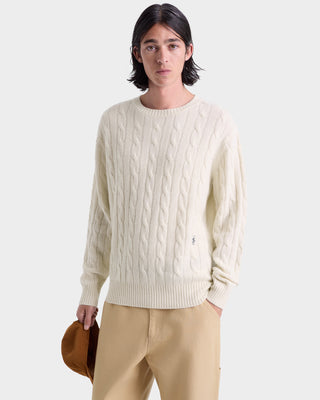 Person wearing a cream cable knit sweater and beige pants holding a brown bag on a white background