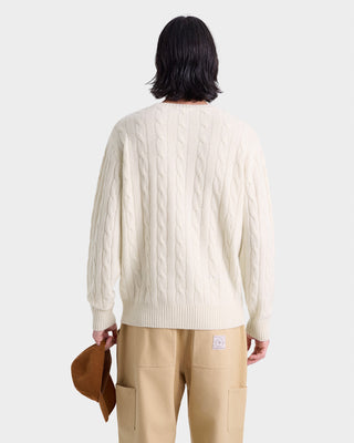 Person wearing a cream cable knit sweater and beige pants holding a brown jacket on a white background