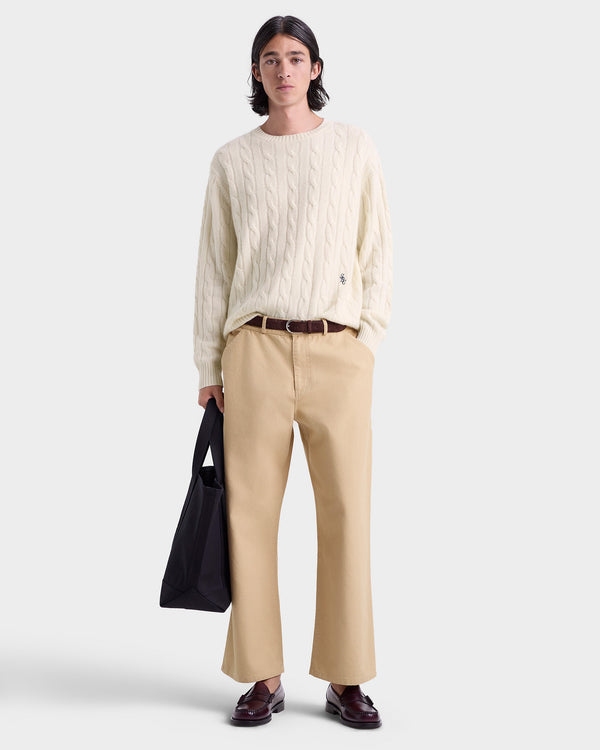 Person wearing a cream sweater and beige pants holding a black bag on a white background