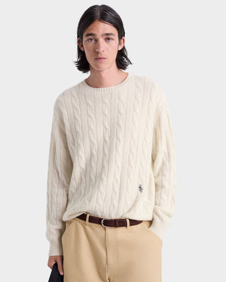 Person wearing a cream cable knit sweater and beige pants on a white background