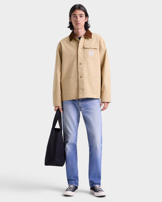Person wearing a beige jacket, blue jeans, and black shoes on a white background