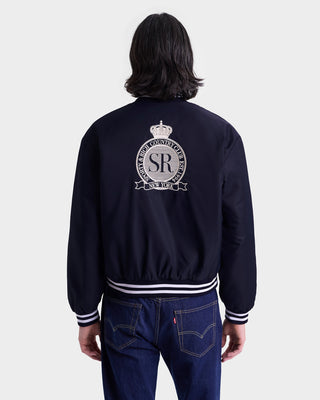 A person in a Royal Club Nylon Varsity Jacket - Navy/White with a crest and SR logo on the back, paired with blue jeans, is facing away.