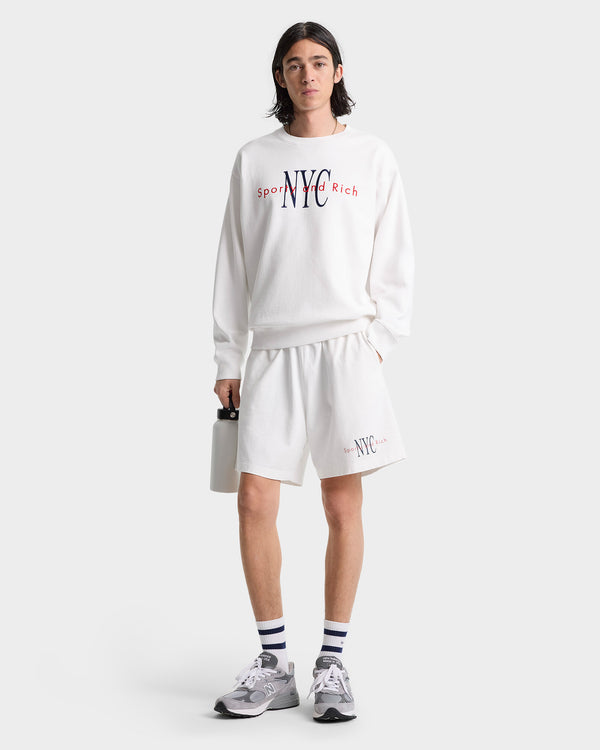 Person wearing a white NYC sweatshirt and NY Minute Gym Short - White/Navy, holding a water bottle, standing against a plain background.