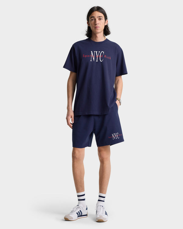 Man wearing a navy NYC t-shirt, NY Minute Gym Short in navy/white with a drawstring waistband, striped socks, and white sneakers, standing against a white background.