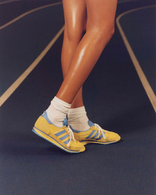 Wearing white socks and the retro-runner styled Adidas SL72 - Yellow/Blue sneakers, an individual strides confidently on a running track.