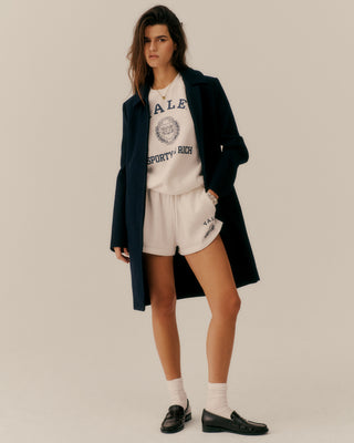 Woman wearing a dark coat over a white t-shirt with text, white shorts, and black shoes on a plain background