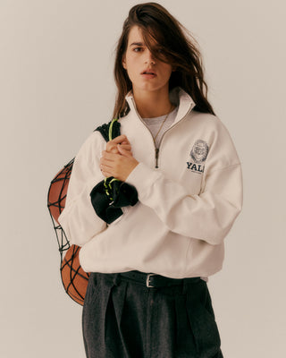Person wearing a white quarter zip with a logo, holding a basketball, on a plain background