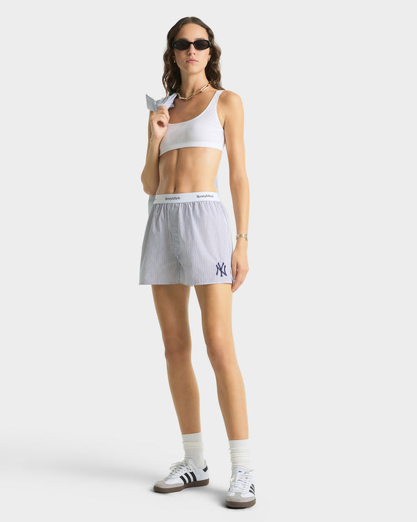 Woman wearing a white tank top and shorts with a logo, holding a small object on a plain background