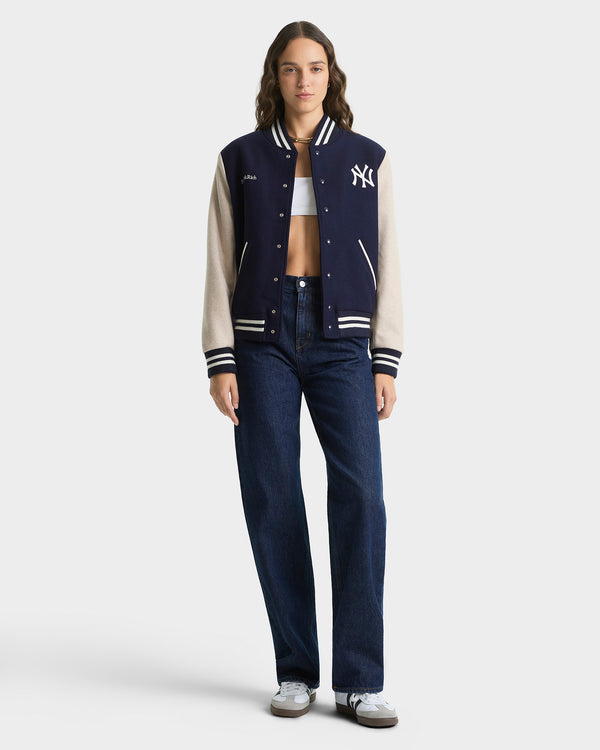 Person wearing a navy and beige varsity jacket with a logo, white shirt, and dark jeans on a white background