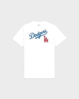A woman on a baseball field wearing a white Los Angeles Dodgers t-shirt from the back, featuring the team's script logo and 'LA' logo in blue and red, paired with blue athletic shorts and a white cap.