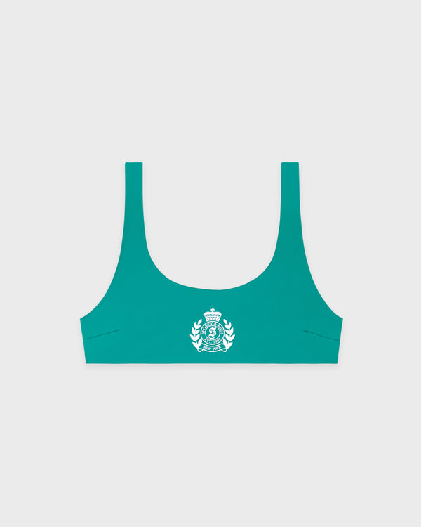 A woman wears the NY Crest Claudia Bikini Top in Amalfi Green/White, featuring a metal clasp closure and crafted from ECONYL regenerated nylon, as she stands against a plain white background facing forward with a neutral expression.