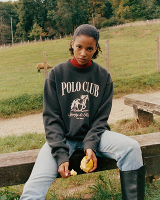 Person wearing a Polo Club sweatshirt sitting on a wooden fence outdoors.
