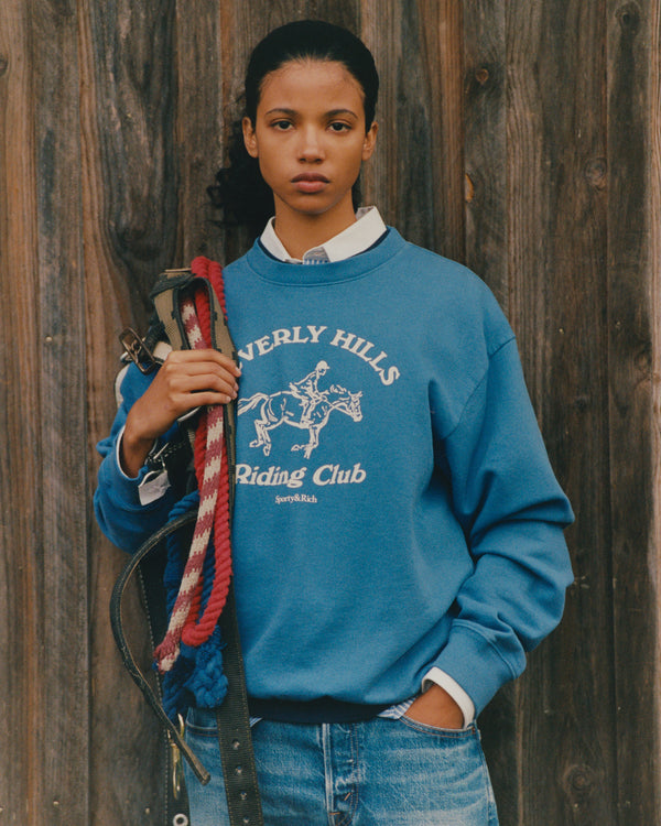 Person wearing a blue 'Everly Hills Riding Club' sweatshirt holding horse reins against a wooden background