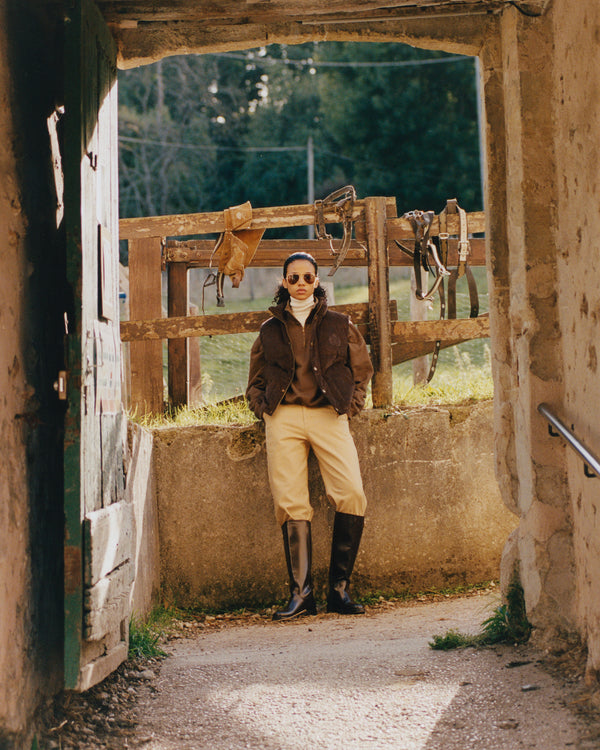 Person in equestrian attire standing in a stable doorway with tack hanging on the wall.