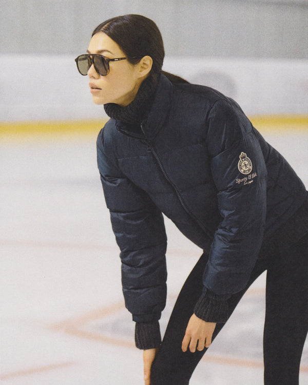 A woman in sunglasses and a dark puffer jacket stands on an ice rink, her SRC Turtleneck - Dark Gray adding sophistication to her winter look.