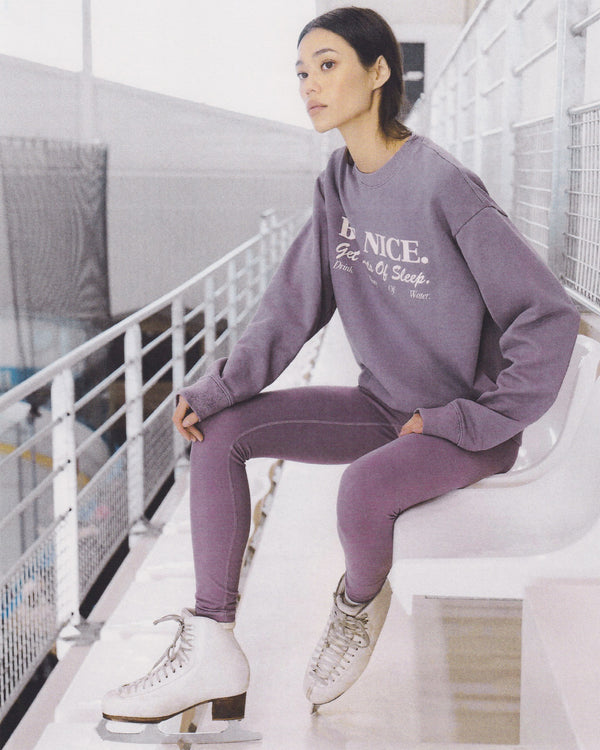 Woman in a purple sweatshirt and SR Bold Legging - Eggplant/White, wearing ice skates, sits on a bench by an indoor ice rink.