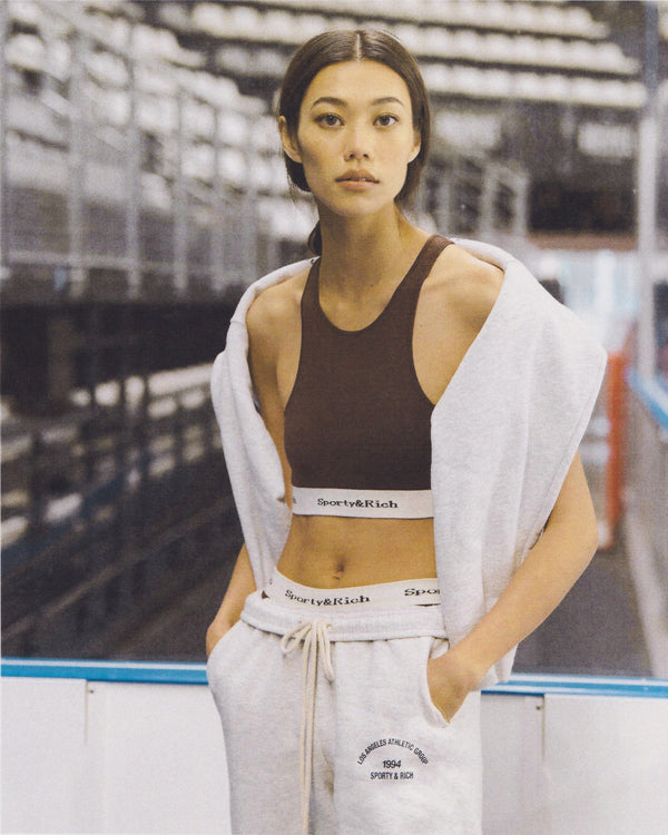 A woman stands in an indoor arena sporting the Serif Logo Knitted Cropped Tank in Chocolate/Off White, styled with loungewear.