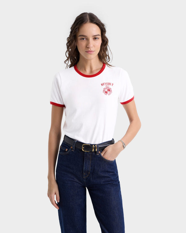 A woman wearing the Beverly Hills Crest Soft Ringer Tee in White/Ruby, made from 100% cotton, pairs it with dark jeans and has her hand in her pocket against a gray backdrop.