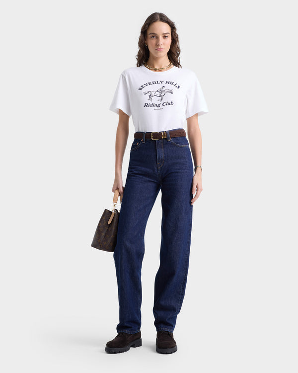 A woman wearing the Beverly Hills Riding Club Soft T-Shirt in White/Navy and jeans holds a handbag against a plain backdrop. Crafted in Portugal, this 100% cotton shirt combines style with comfort seamlessly.