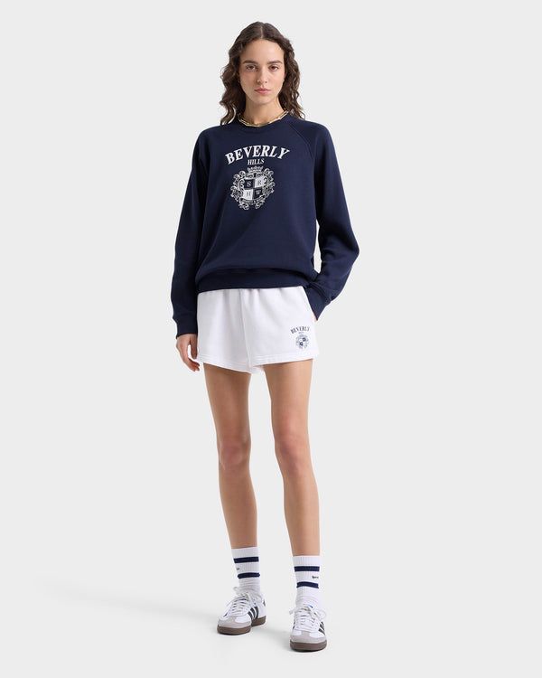 A woman in a navy sweatshirt and white Beverly Hills Crest Soft Disco Shorts with an elastic waistband stands with hands in pockets, wearing striped socks and sneakers.