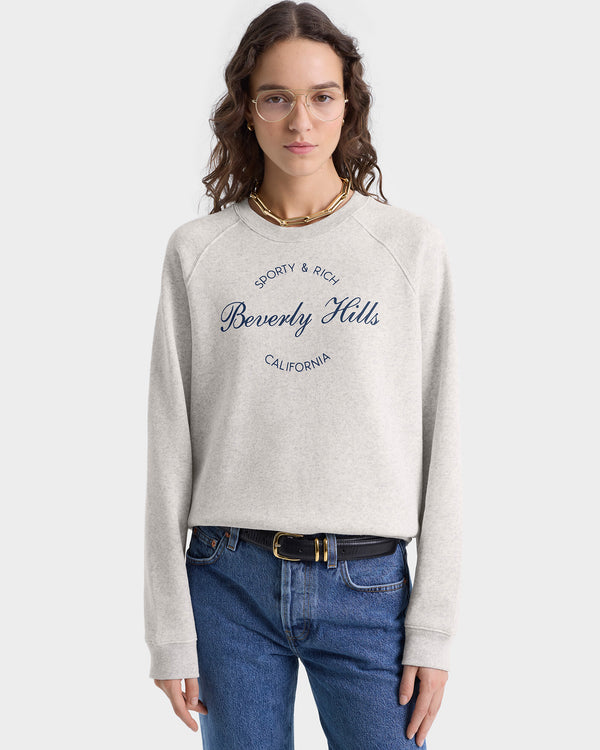 A woman in glasses and jeans stands against a plain background, wearing the Beverly Script Soft Crewneck in Heather Gray/Navy.