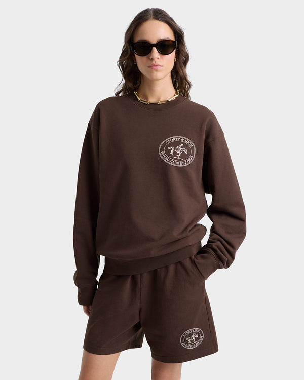 Wearing the Riding Club Crewneck in Chocolate/Cream with matching shorts, this person sports a 100% cotton outfit featuring a circular logo. Made in the USA and resistant to shrinking, the ensemble against a plain background is completed with sunglasses.