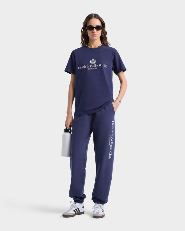 A person in sunglasses, dressed stylishly in a Health & Wellness Club outfit, pairs it with H&W Crest Sweatpant - Navy/White. They hold a water bottle, embodying both fashion and fitness chic.