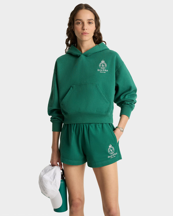 Person wearing a green hoodie and shorts set with a logo, holding a white and green bag.