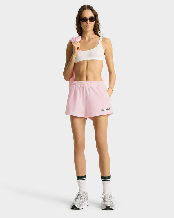 Woman in sunglasses wears a white sports bra, Syracuse Embroidered Disco short - Pink Dolphin/Navy with elastic waistband, white sneakers, and holds a pink sweatshirt. Made in Portugal.