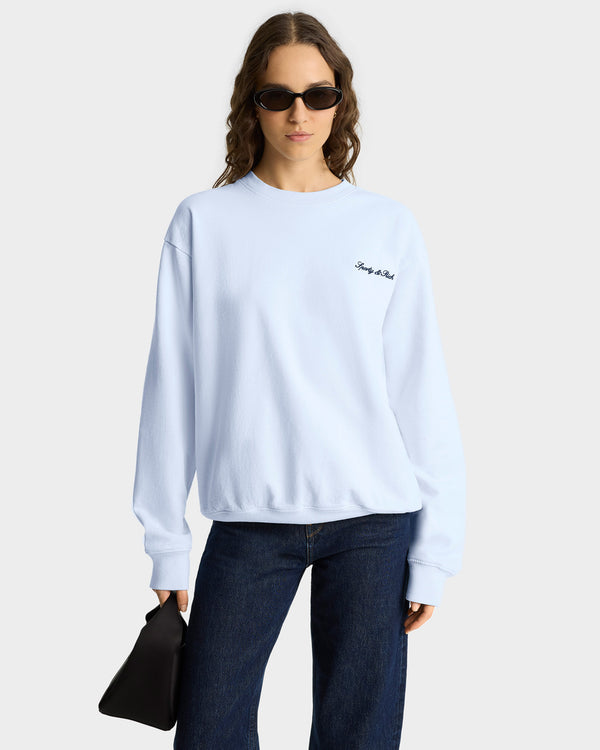 A woman in sunglasses wears the Syracuse Embroidered Crewneck - Ciel/Navy, made from USA-grown cotton, paired with dark jeans and a black clutch against a plain background.