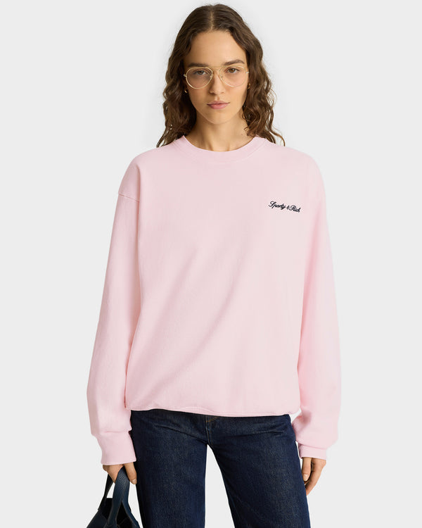 Woman wearing a Syracuse Embroidered Crewneck - Pink Dolphin/Navy sweatshirt, blue jeans, round glasses, and holding a black bag.
