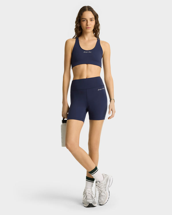 Woman in Syracuse Biker Shorts - Navy/White and sports bra, holding a water bottle, paired with white sneakers and striped socks—ideal luxe activewear for style-conscious fitness fans.