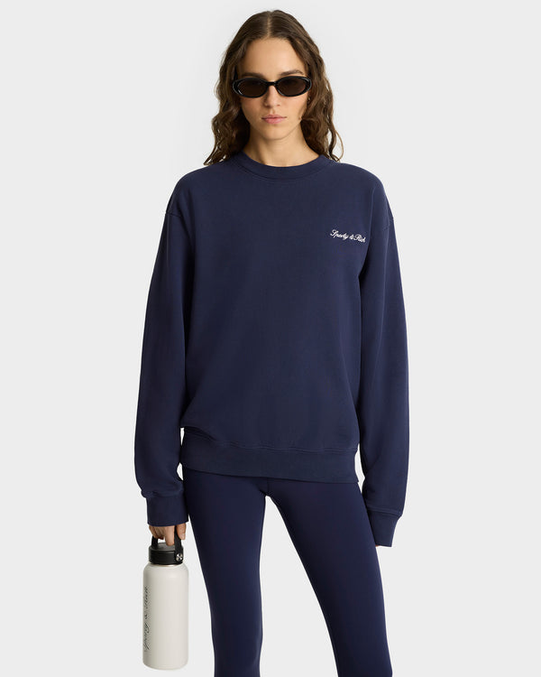 Woman wearing the Syracuse Embroidered Crewneck in Navy/White, made from USA grown cotton, paired with leggings and sunglasses, holding a white water bottle against a plain background.