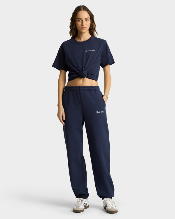 A woman stands facing forward in navy Syracuse Embroidered Sweatpants and a matching knotted t-shirt, both made from USA-grown cotton, paired with white sneakers.