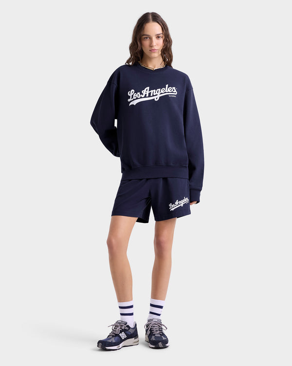Against a plain background, a person wears a navy Los Angeles sweatshirt paired with Dodgers Gym Short - Navy/White, featuring an elastic waistband. The outfit is completed with sneakers and striped socks, highlighting comfort and style in 100% cotton fabric.